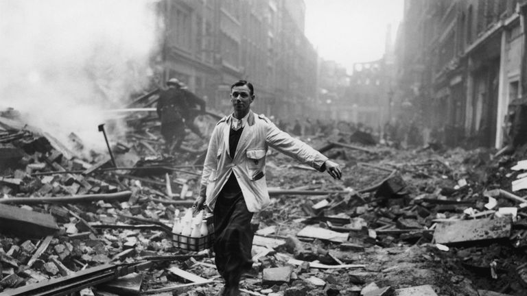 A milkman delivering milk in a London street devastated during a German bombing raid. Firemen are dampening down the ruins behind him