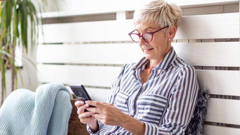 Mature woman with mobile phone on her hands sitting in room and sending messages to her friends and family